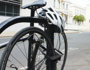 Bike locked up in a town to a metal bike rack with a D lock securing the rear wheel and frame and the security cable securing a helmet