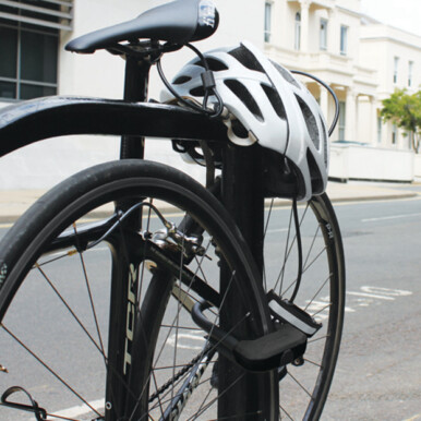 Bike locked up in a town to a metal bike rack with a D lock securing the rear wheel and frame and the security cable securing a helmet